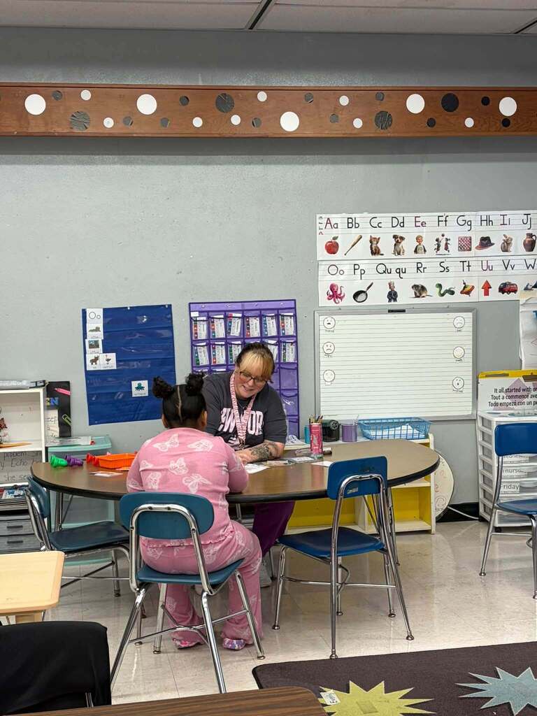 A classroom scene shows an adult seated across a table from a child, working together on papers during one-on-one instruction. The room features educational displays including an alphabet chart showing "Aa, Bb, Cc, Dd, Ee, Ff, Gg, Hh, Ii, Jj; Oo, Pp, Qu qu, Rr, Ss, Tt, Uu, Vv, Ww" and a partial multilingual sign reading "It all started with…," "Tout a commencé av…," and "Todo empezó con un p…" in English, French, and Spanish. The classroom includes picture charts, organized storage, chairs, and a whiteboard with ruled lines and small face drawings, creating a structured learning environment that supports individualized literacy instruction and celebrates linguistic diversity.