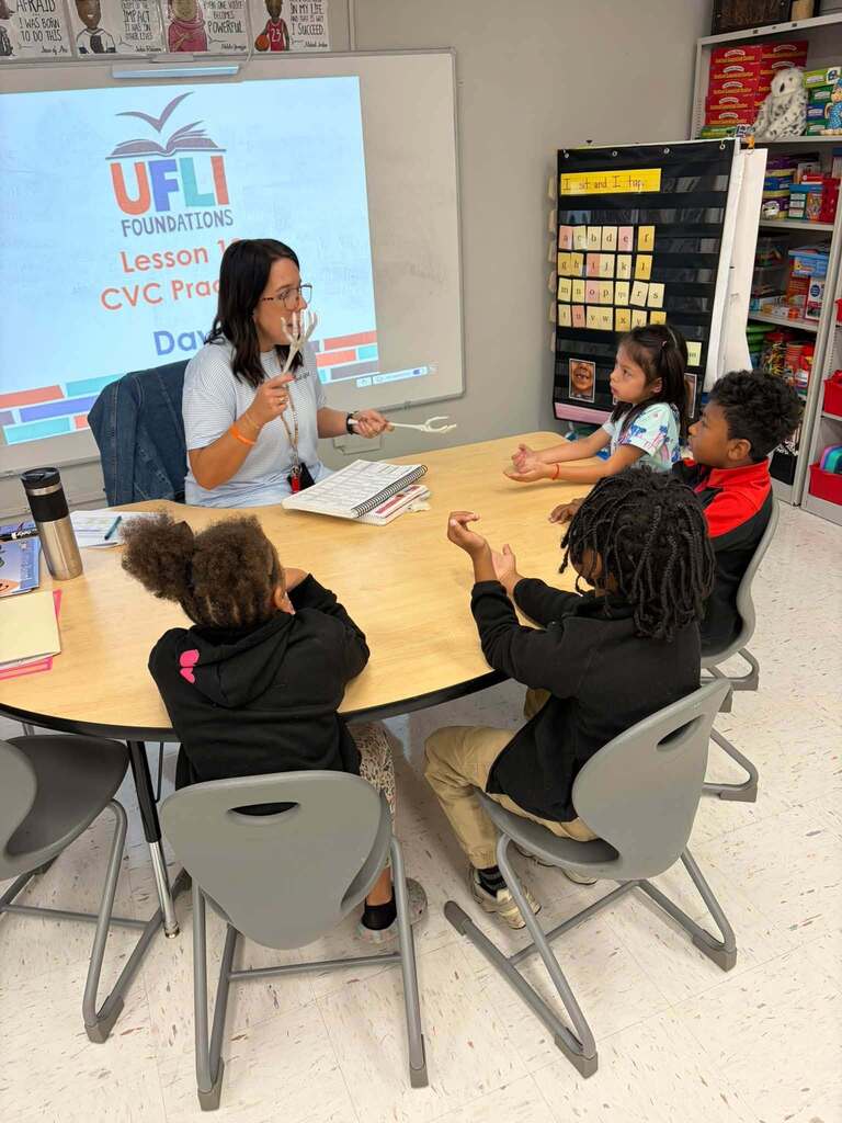 A teacher leads a small-group phonics lesson at a classroom table. Several children watch and participate in the interactive lesson. Behind the teacher, a projected presentation displays "UFLI Foundations Lesson 1 CVC Practice Day [unclear]," and a letter-card pocket chart is visible on the wall. The hands-on, multisensory instructional approach using physical models and visual supports demonstrates evidence-based literacy instruction focused on helping students understand how sounds are produced and connected to letters in consonant-vowel-consonant (CVC) word patterns.