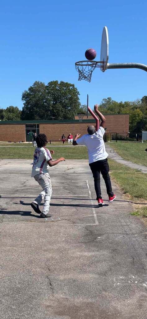 Two people are on an outdoor basketball court as one watches a ball arc toward a hoop in the upper right. A brick school building, trees, and several people are visible in the background.
