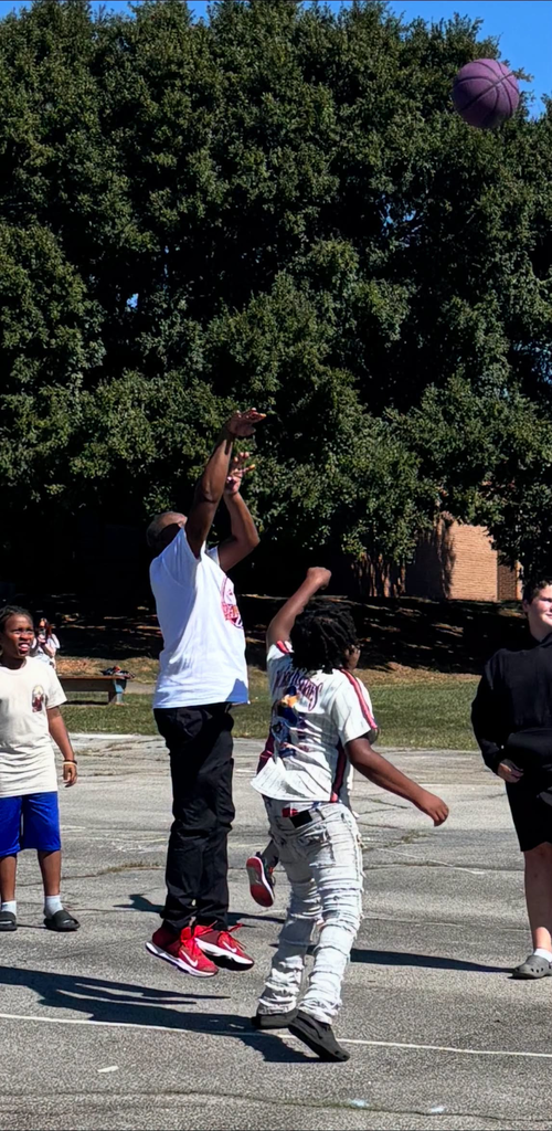 Two people play basketball on an outdoor paved court as one shoots toward the hoop, with the ball just above the rim. A brick school building, grass, trees, and a clear blue sky are visible in the background.