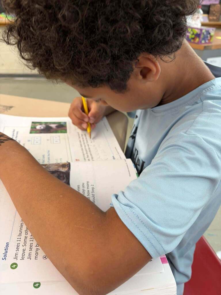 A student in a light-blue shirt leans over a workbook and writes with a yellow pencil. The open pages show math problems and small pictures, though most of the text is obscured or blurred.