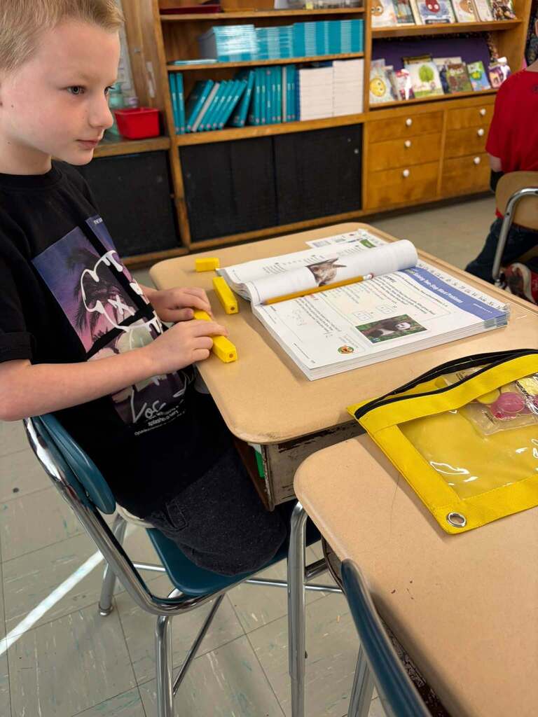 A student handles yellow counting blocks at a classroom desk beside an open workbook and pencil. Books and classroom materials are on shelves in the background, and another student is partly visible on the right.