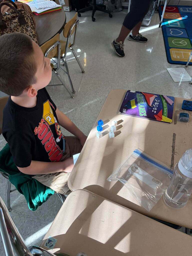 A child sits at a classroom desk with small blue-and-white blocks arranged on the surface. Desks, a backpack, books, a water bottle, and a colorful floor mat are visible around the classroom.