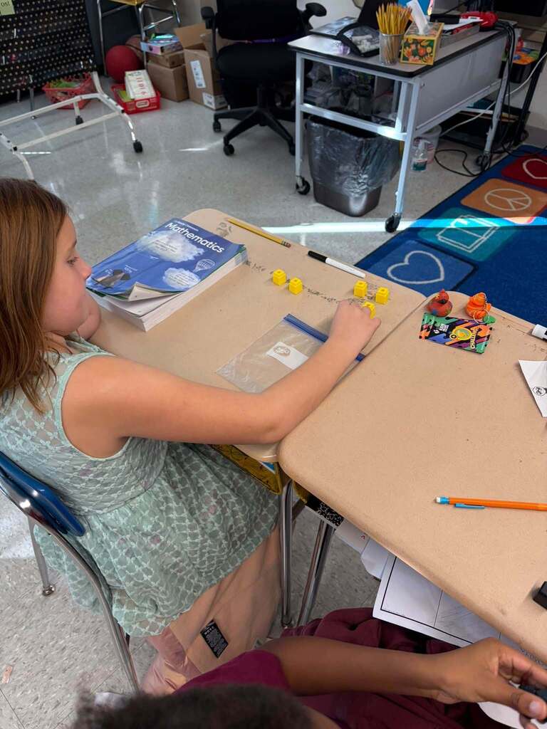 A student works with small yellow counting cubes at a classroom table beside a textbook labeled “Mathematics” and other materials. Desks, a chair, and a colorful rug are visible in the background.