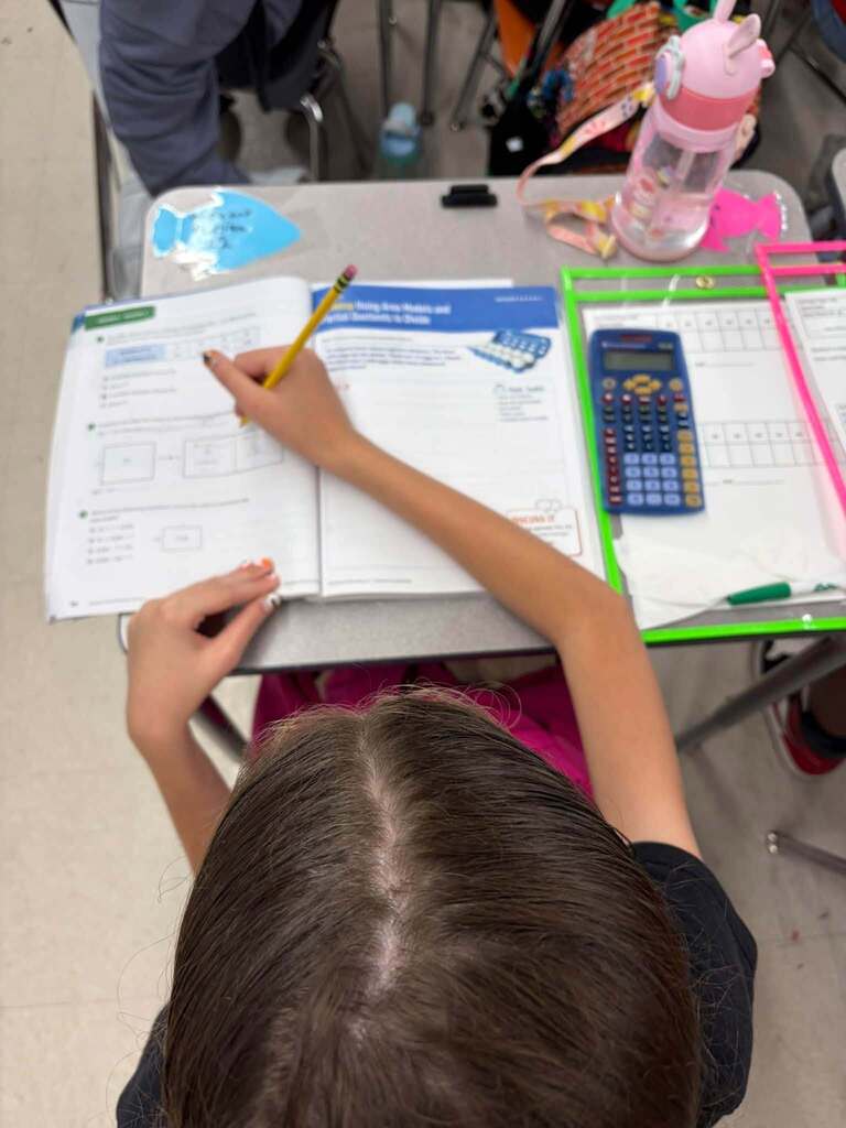 A student writes in an open workbook with a pencil at a classroom desk. A calculator, worksheets, and a pink water bottle are on the desk.