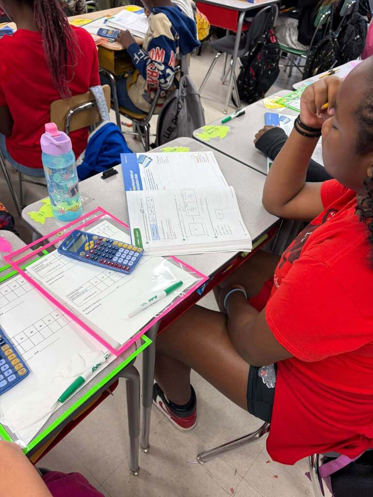 Several students sit at classroom desks working on math worksheets. The desks hold open workbooks, laminated worksheets, calculators, markers, and a water bottle; backpacks hang from chairs in the background.