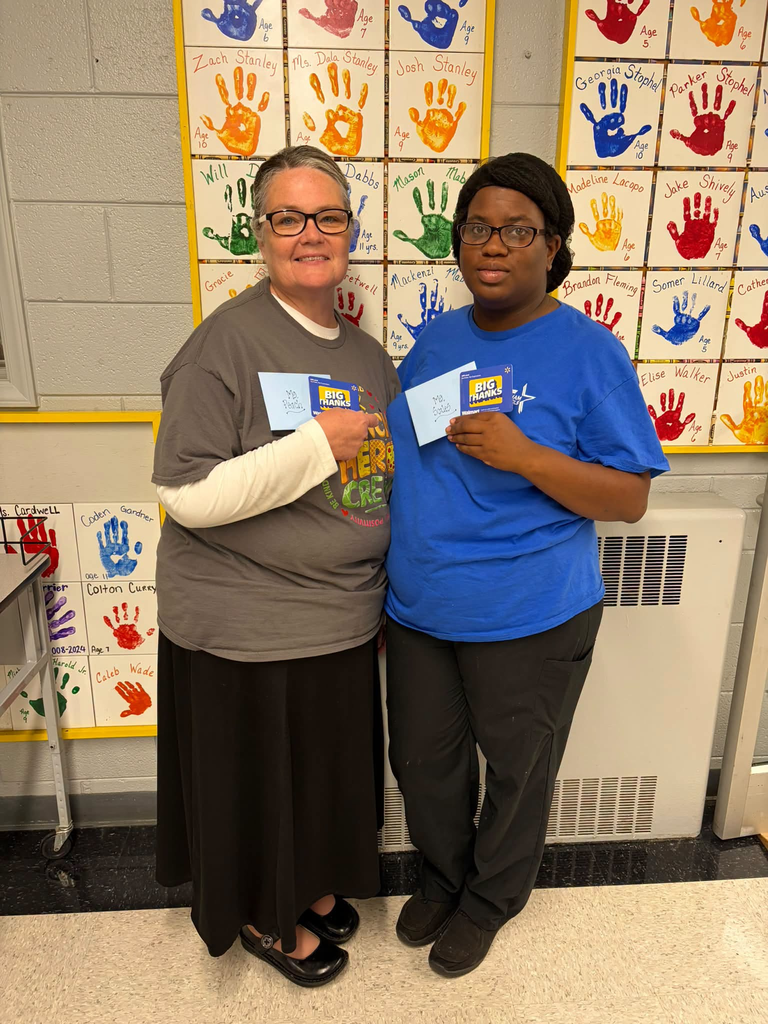 Two people stand side by side indoors, each holding a small blue card with a yellow-and-blue logo. Behind them, colorful classroom displays feature handprints with names written beside them.