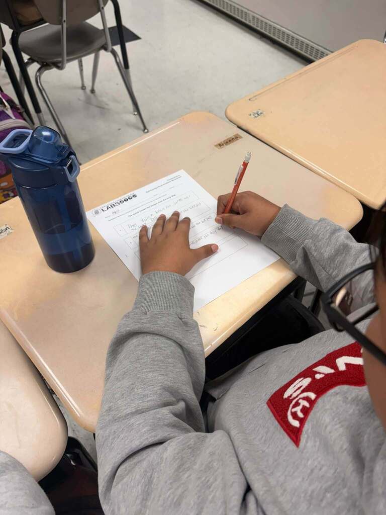 A student in a gray sweatshirt writes with a pencil on a worksheet at a classroom desk. A blue water bottle sits beside the paper, with classroom chairs and another desk nearby.