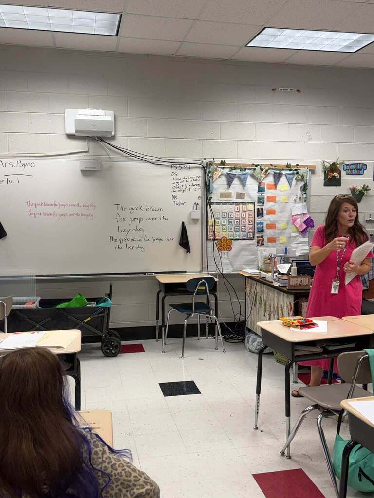 A teacher stands beside classroom desks holding papers. The whiteboard displays handwriting practice sentences reading “The quick brown fox jumps over the lazy dog” and asks, “How do we use a grid to show exactly where an object is?” A wall calendar and a bulletin board decorated with bunting and student materials are also visible.