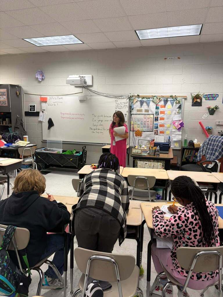 A teacher in a bright pink dress stands at the front of a classroom holding papers while students sit at desks and write. The whiteboard displays the word “Vocabulary,” and classroom displays, bookshelves, and supplies are visible around the room.