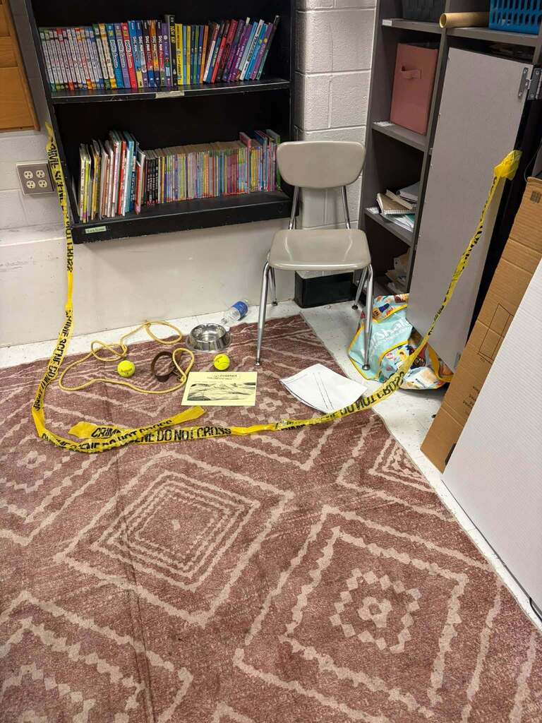 A classroom or storage area is staged like a crime scene, with yellow “CRIME SCENE DO NOT CROSS” tape surrounding objects on a patterned rug. The scene includes a chair, metal bowl, blue bottle, yellow balls, loop of rope, dark curved object, printed sheet, and loose white page; bookshelves and storage cabinets stand in the background.