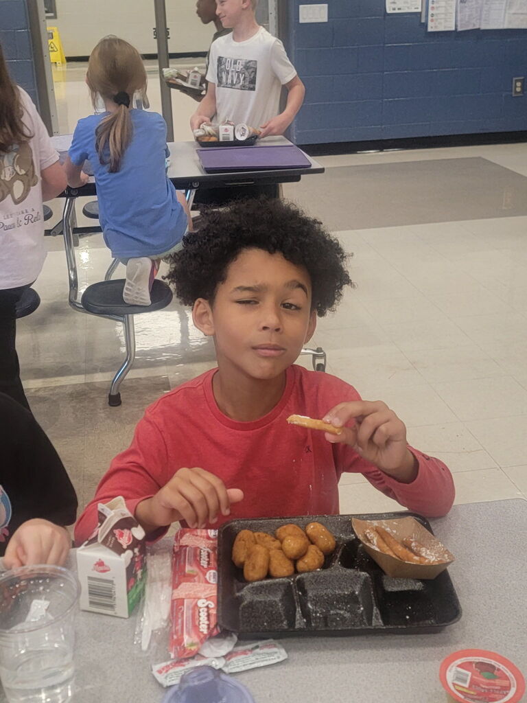 National School Lunch Week 2026- Little boy in long-sleeved red shirt making a winking face while eating chicken nuggets, fried dessert covered in sugar powder, and chocolate milk