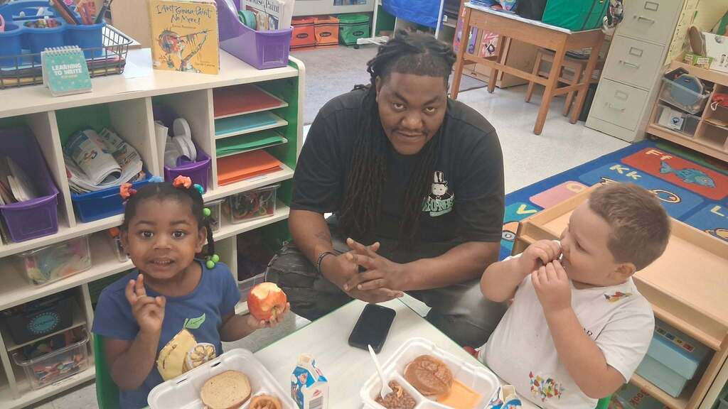 Father (middle) sits with young female (left) and male (right) students at table inside classroom while eating lunch