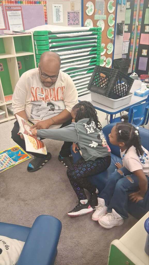 A father (left) reading to two young children while one child (middle) is pointing to something in the book and the other child (right) is looking at the illustrations in the book from a couch inside a classroom