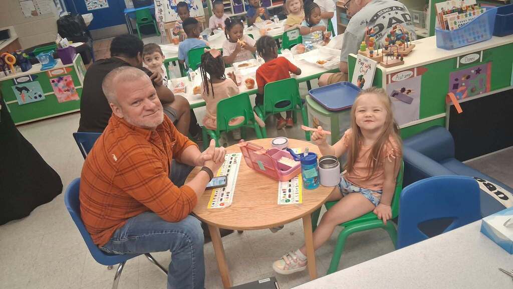 Father (left) sits with young female student (right) at a table inside a classroom while doing math activity