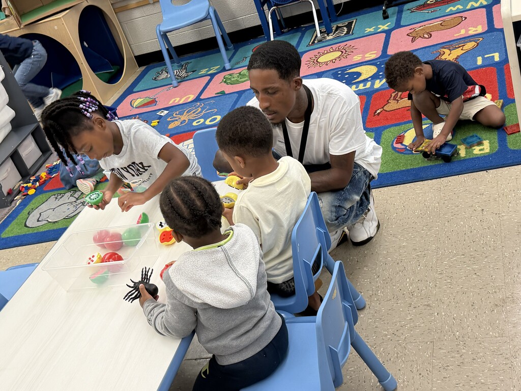 Four young students and a father inside a classroom. Three students are at a table with the father while another is in the background on the floor playing with a toy