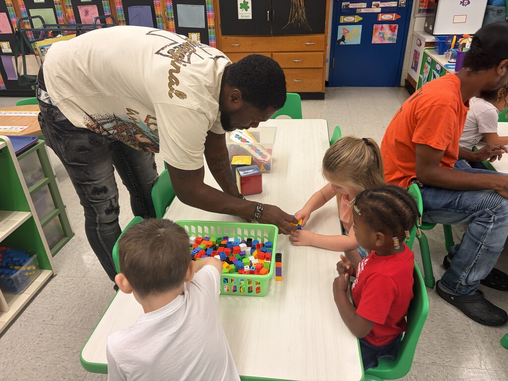 Three students and a father at a table inside a classroom using colorful building blocks during a math activity