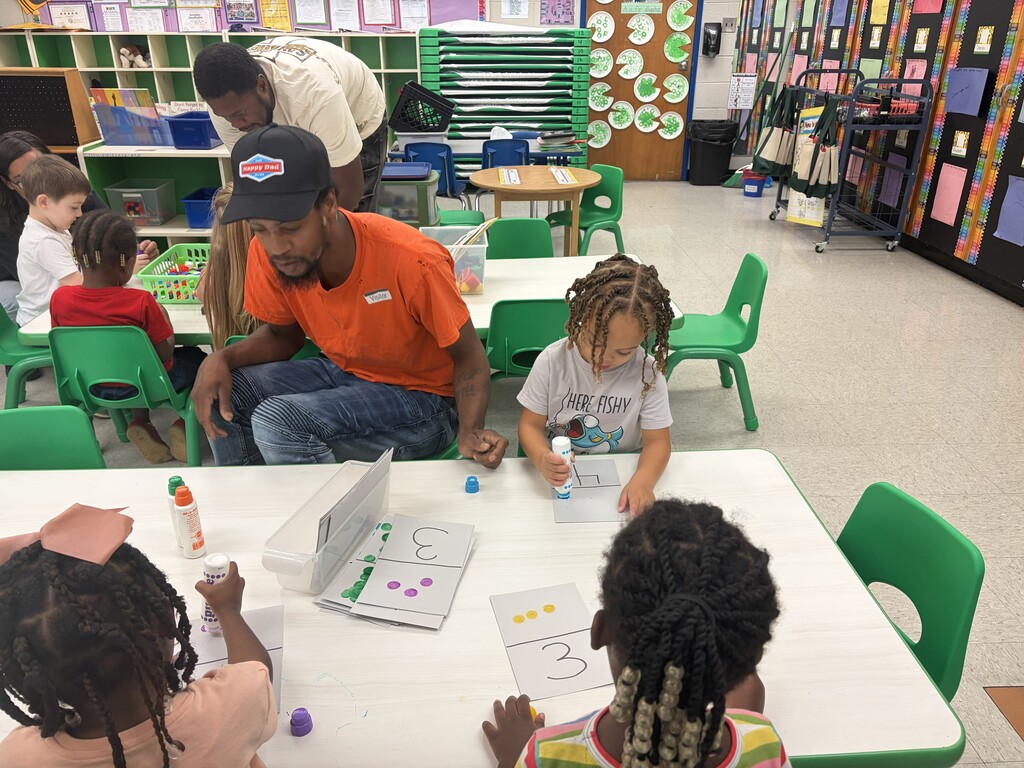 Two tables inside classroom- One in the back with three young students and a father helping with a math activity and the front table with three young students and a father helping with a numbers activity