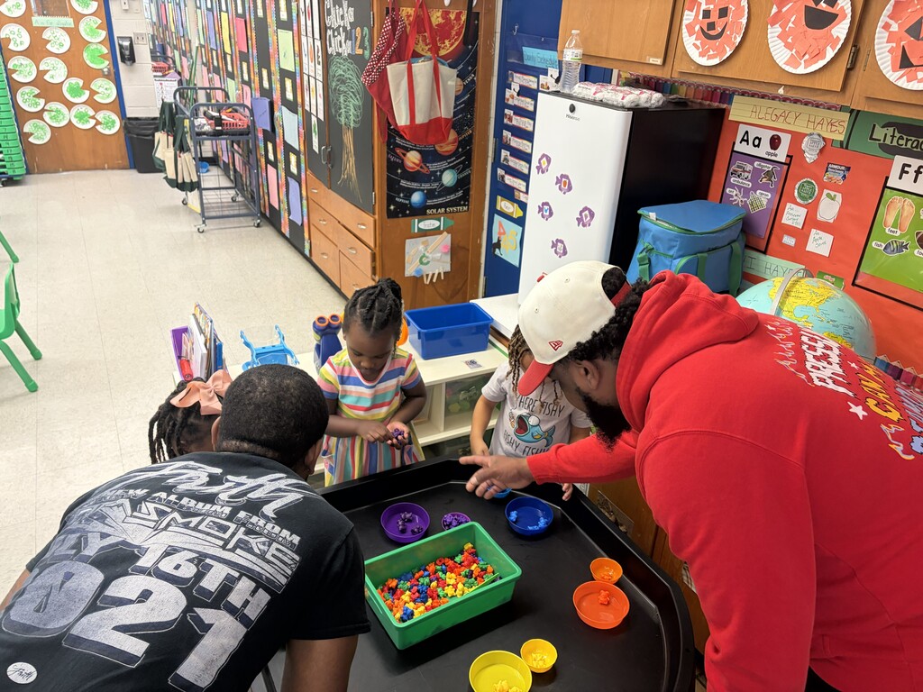 Three students and two fathers inside a classroom at a table with colorful blocks and bowls and engaging in a math activity
