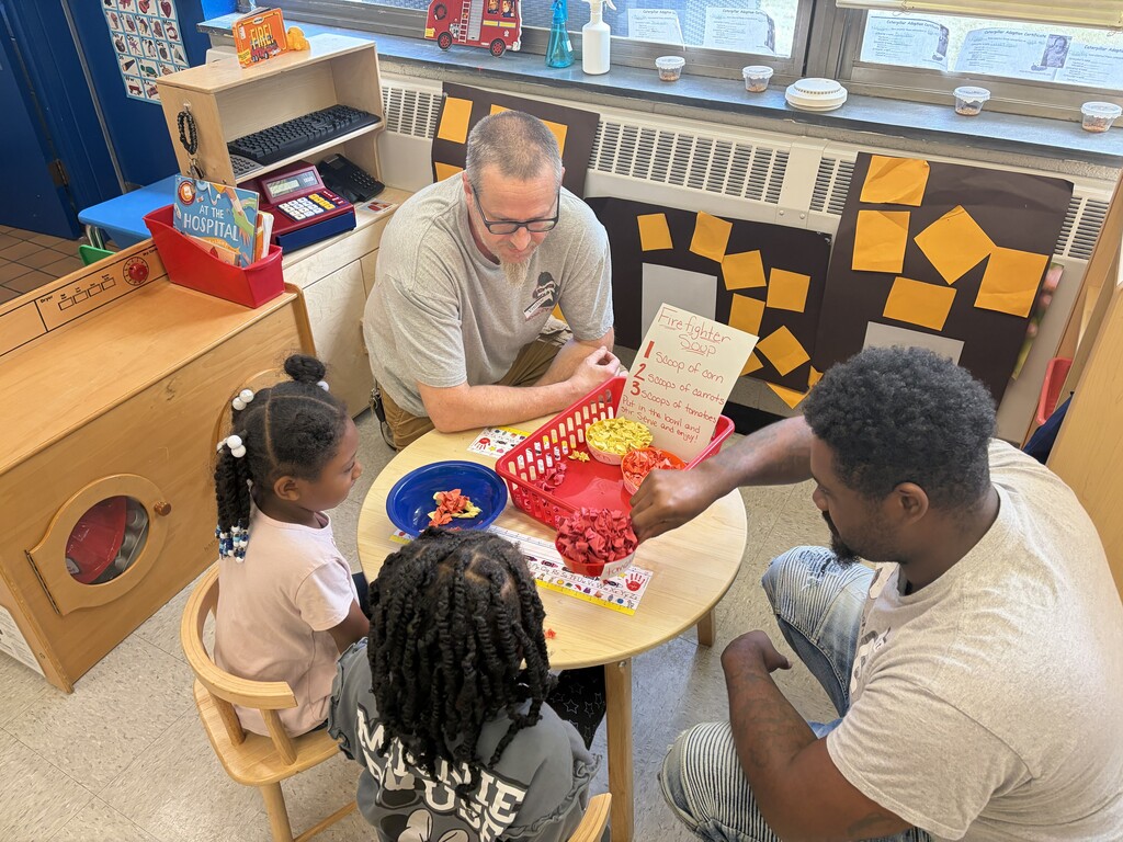 Two young students (bottom left) sit at a table inside a classroom with two fathers (top left and right side) at a table inside a classroom while doing a math activity