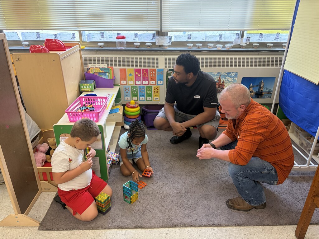 Two young children (left) on the floor using building materials and two fathers (right) observing them inside a classroom