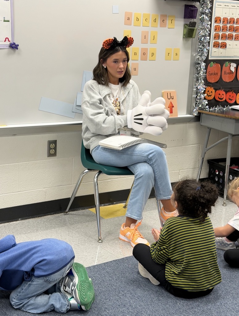 Red Ribbon Week! Lincoln Elementary School- A teacher is sitting in the front of her classroom wearing Mickey Mouse stuffed hands and Halloween Disney ears. There are three students sat on the floor in front of her
