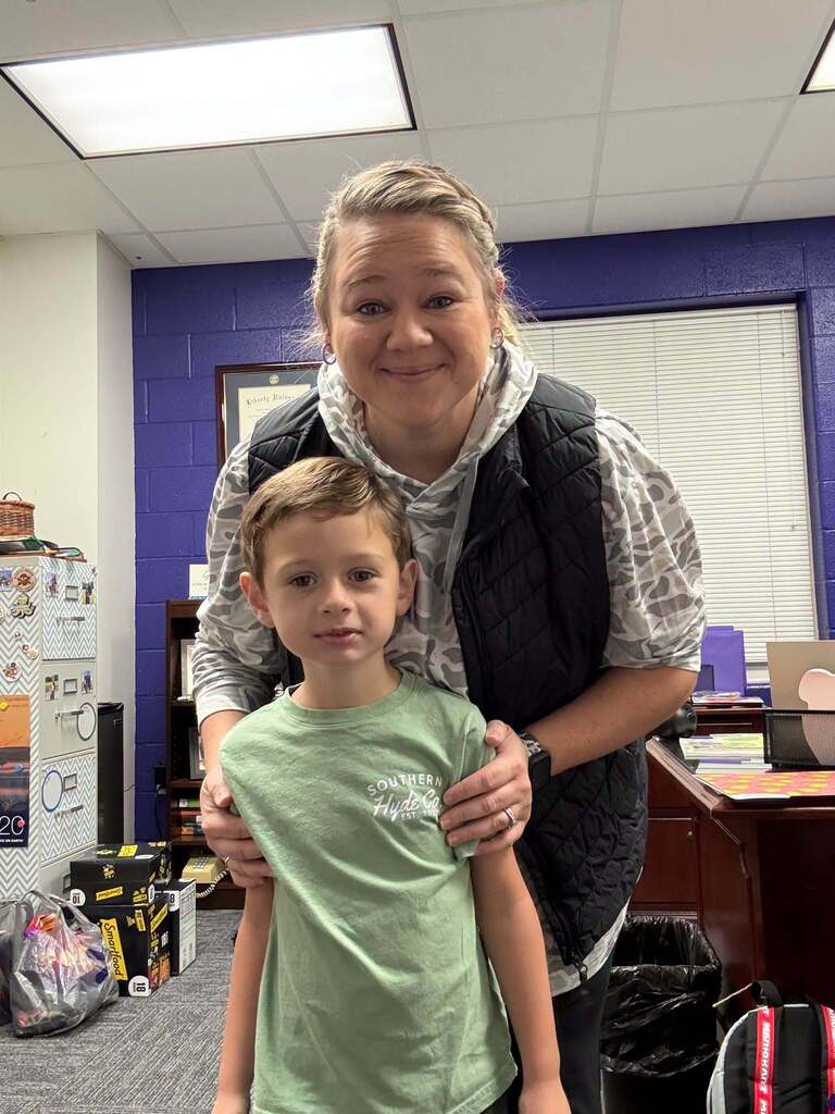 Red Ribbon Week- Lincoln Elementary Camo Day: A teacher standing behind her student. teacher is wearing a black puffer vest with a gray camo t-shirt. the student is wearing a green hunting brand t-shirt