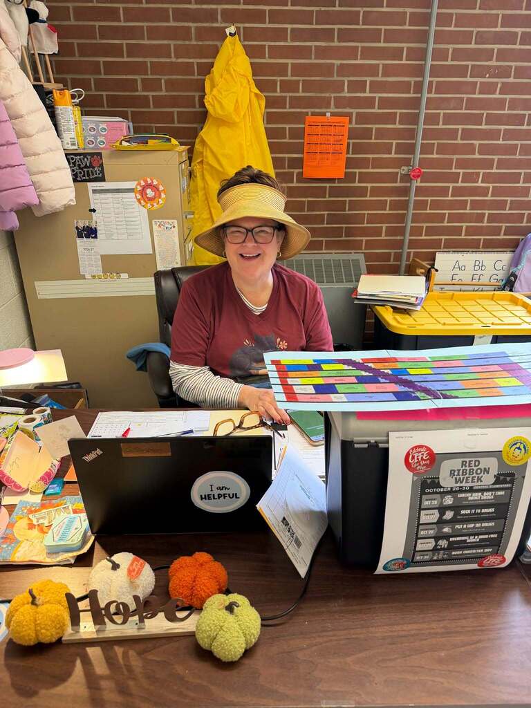Red Ribbon Week- Central Elementary Hat Day: A teacher at her desk wearing a sun hat that has an opening on the top