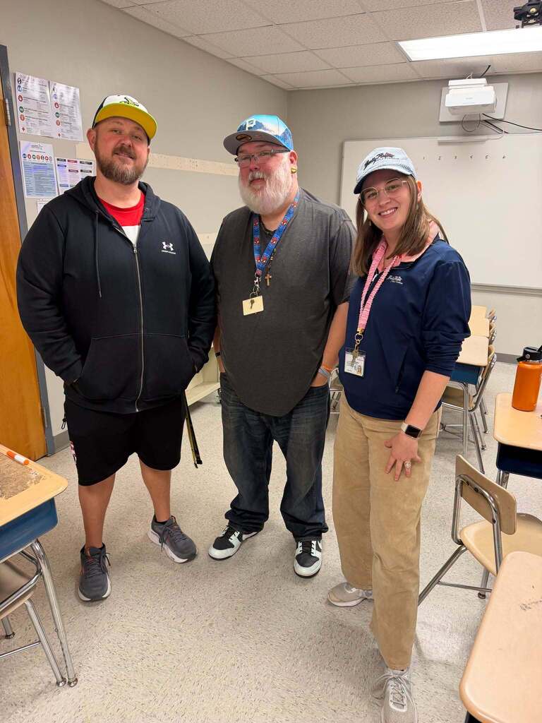 Red Ribbon Week- Central Elementary Hat Day: 3 teachers standing in a classroom (2 men, 1 woman). Left is wearing a white, green and yellow hat. Middle is wearing a blue hat, and right is wearing a white hat with dark wording on the front