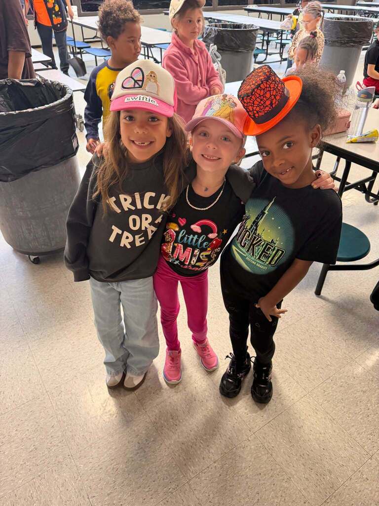 Red Ribbon Week- Central Elementary Hat Day: 3 little girls in the lunch room. Left is wearing a white and pink hat with a heart/peace sign symbol, middle is wearing a pink hat with characters on it, right is wearing an orange Halloween top hat with a black spider web design
