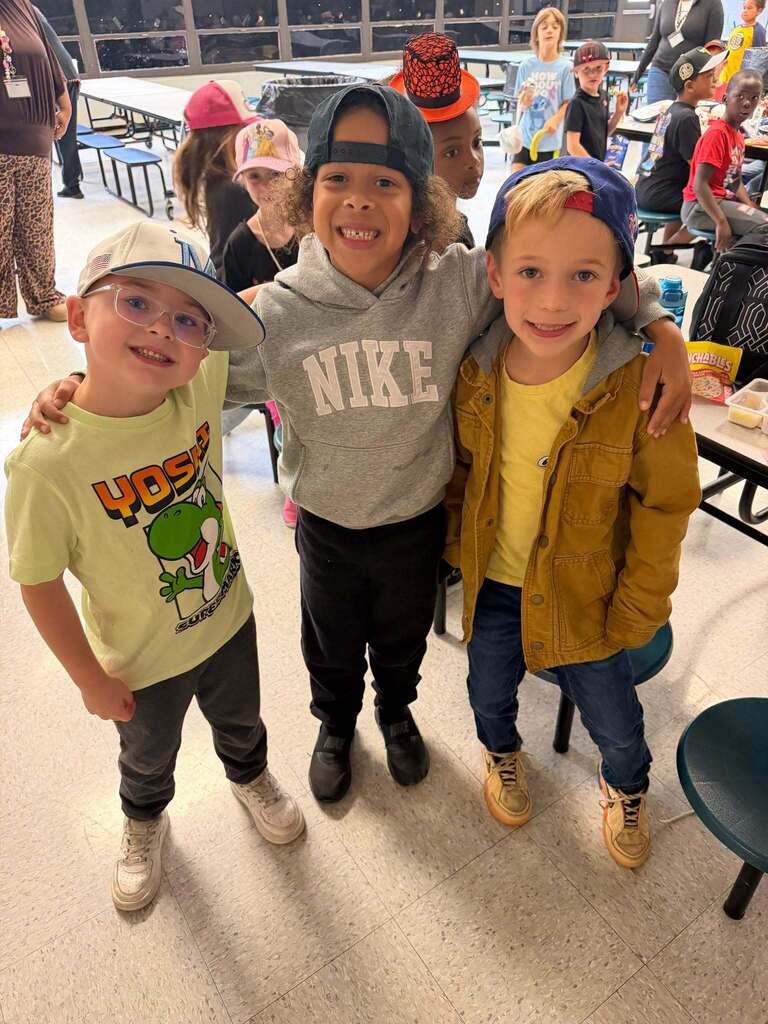 Red Ribbon Week- Central Elementary Hat Day: 3 little boys in a cafeteria. Left is wearing a white hat with a blue "M" on it, middle is wearing a blue jean hat backwards, and right is wearing a blue and red hat backwards