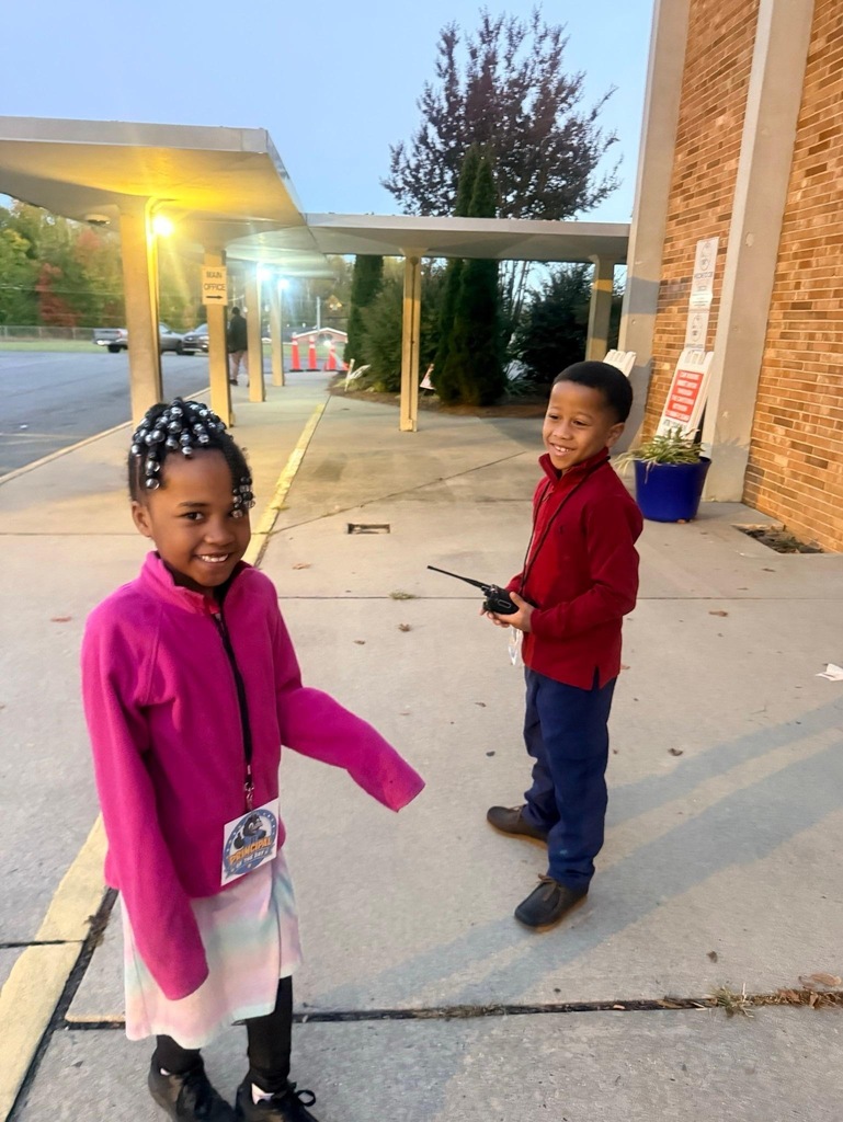 Reidsville Elementary School Principals for a Day- Outside: Little girl on the left wearing a pink pullover on top of a rainbow colored dress and wearing a lanyard/name tag is standing next to a little boy with a red pullover and jeans wearing a lanyard/name tag and holding a walkie talkie