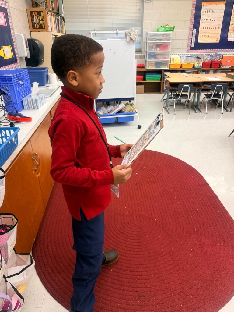 Reidsville Elementary School Principals for a Day- Little boy in red pullover and jeans with a lanyard/name tag holds a clipboard while standing in front of a classroom