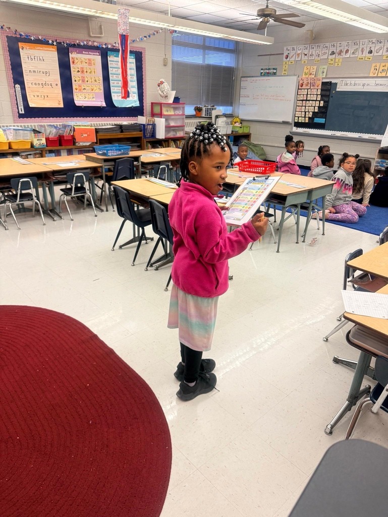 Reidsville Elementary School Principals for a Day- Little girl in pink pullover and rainbow skirt with a lanyard/name tag holds a clipboard while standing in front of a classroom