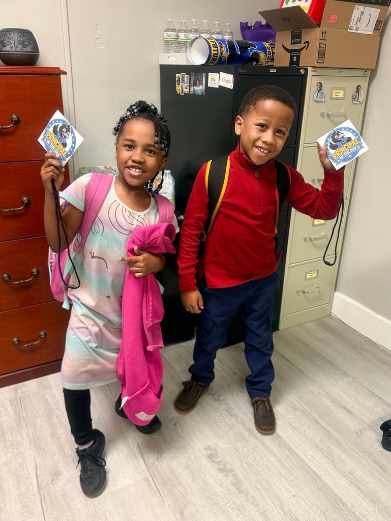 Reidsville Elementary School Principals for a Day- Little girl on the left wearing a unicorn, rainbow dress and holding a pink pullover and name tag standing next to a little boy in a red pullover and jeans who is holding a name tag. Both are smiling