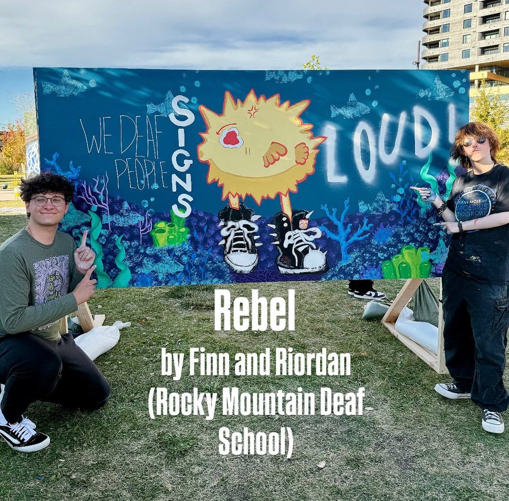 Two students stand outdoors beside a large, colorful mural on an easel. The mural shows an underwater scene with blue water, coral, and fish. At the center is a bright yellow pufferfish wearing black sneakers and shouting. The painted text on the mural reads: “WE DEAF PEOPLE SIGNS LOUD!” The students are smiling and pointing toward the artwork. White overlaid text at the bottom of the photo reads: “Rebel by Finn and Riordan (Rocky Mountain Deaf School).” The setting is grassy with buildings and trees in the background.