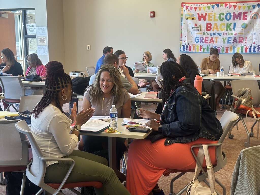 Several people in a classroom sit at tables and chat with each other. A banner hangs on the wall behind them.