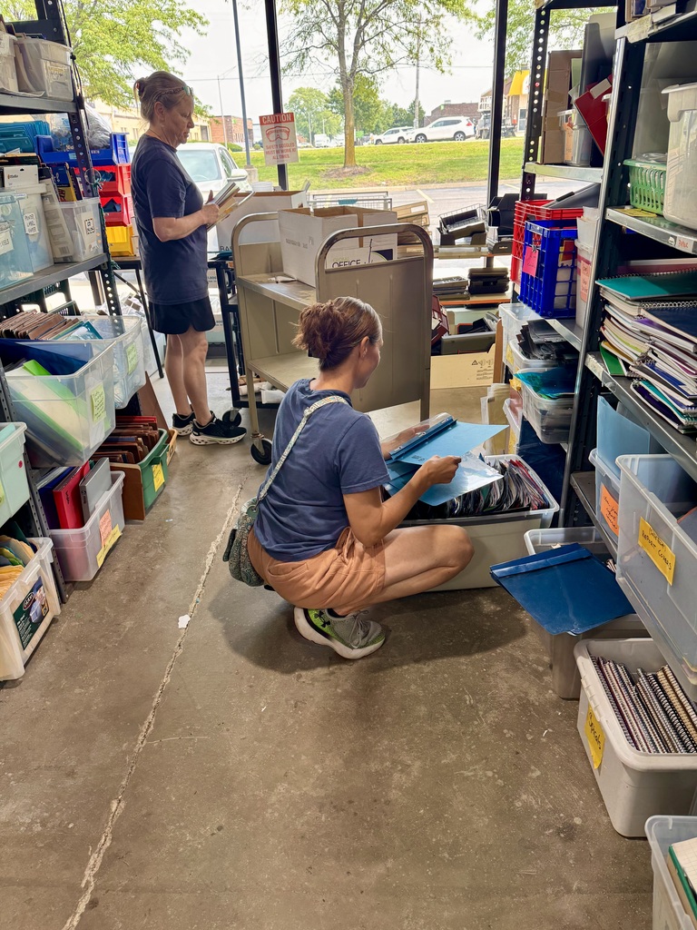 Two people inside a storage space surrounded by shelves and boxes. One is reading, the other is kneeling.