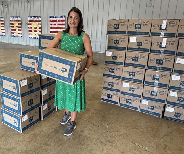 A woman in a green dress holds a box among stacked boxes. A wall behind has flags.