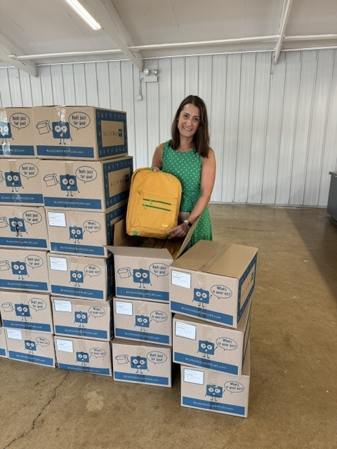 A woman in green holds a yellow backpack and stands before a stack of boxes in a room.