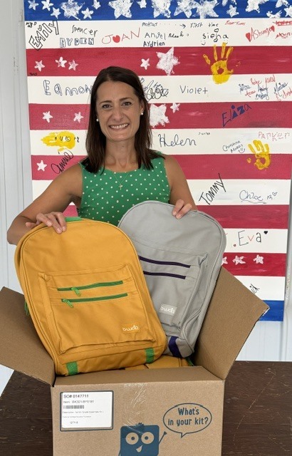 A woman in a green dress holds two backpacks in a box. Behind her, a wall displays a colorful flag with handprints.