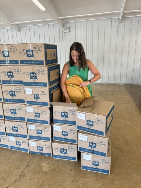 A woman in a green shirt examines a yellow box among a stack of similar boxes in a large room.