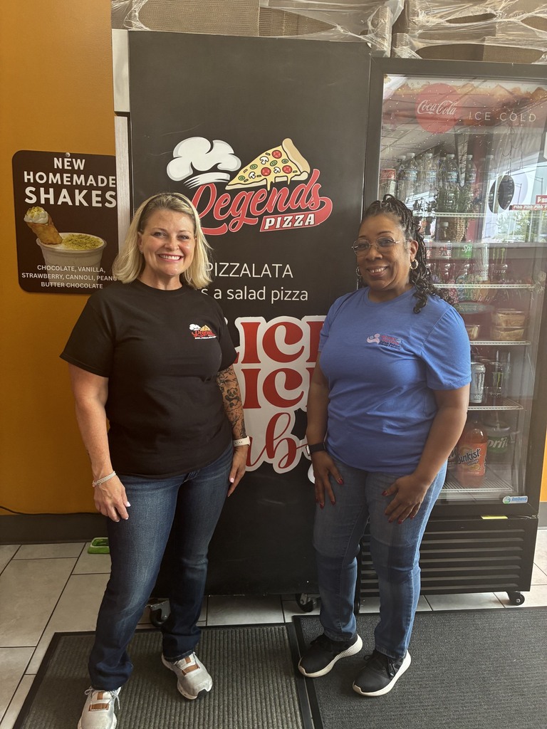Two women stand by a cooler labeled "Legends Pizza" with a salad pizza sign, one in a black shirt, the other in a blue shirt.