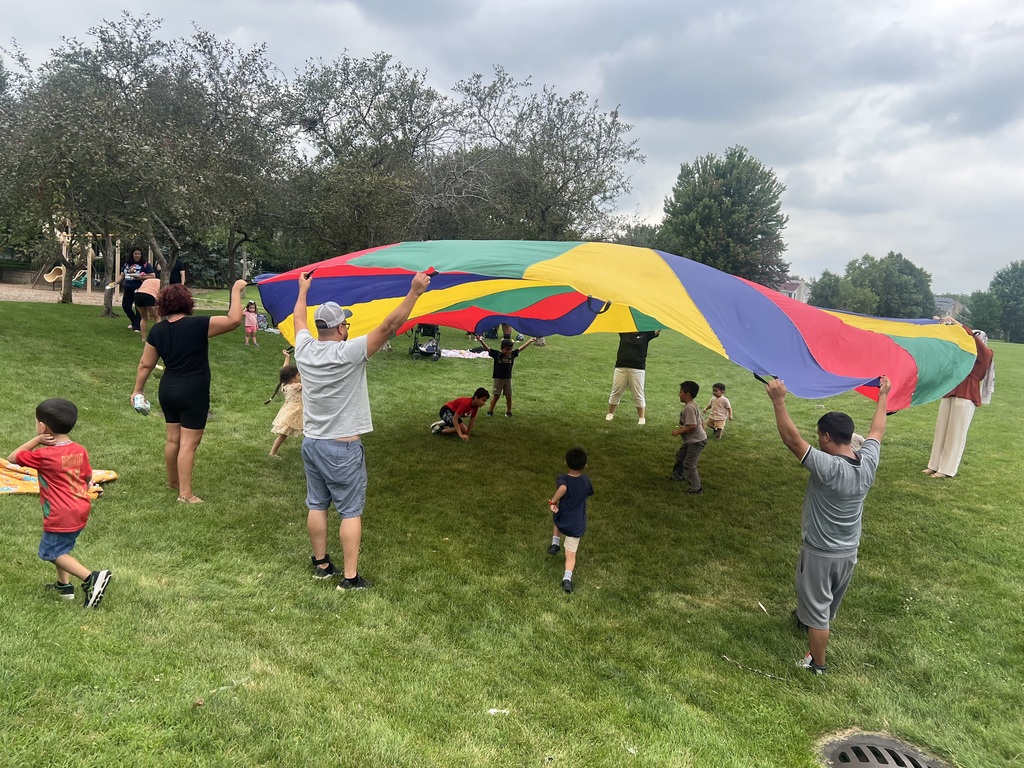 Group of adults and children playing on a grassy field, holding a large multicolored parachute.