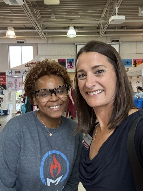 Two women smile inside a room with high ceilings, lights, and a partially visible white tent.