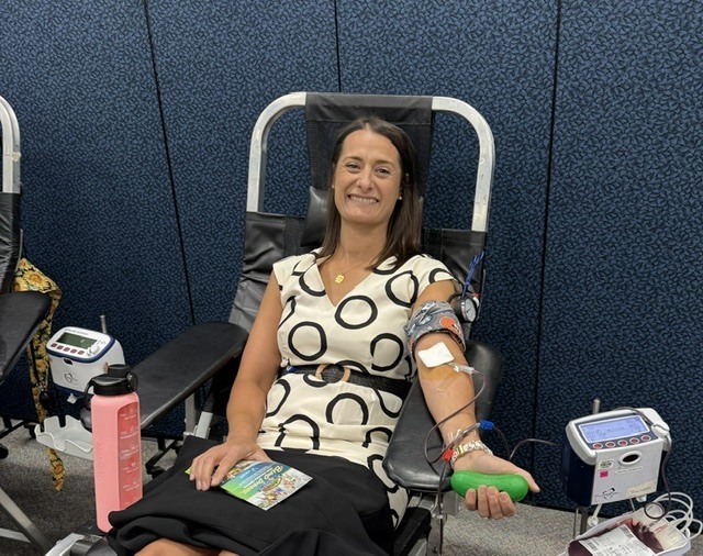 A woman with a blood pressure cuff sits in a chair, smiling. A pink bottle sits next to her.