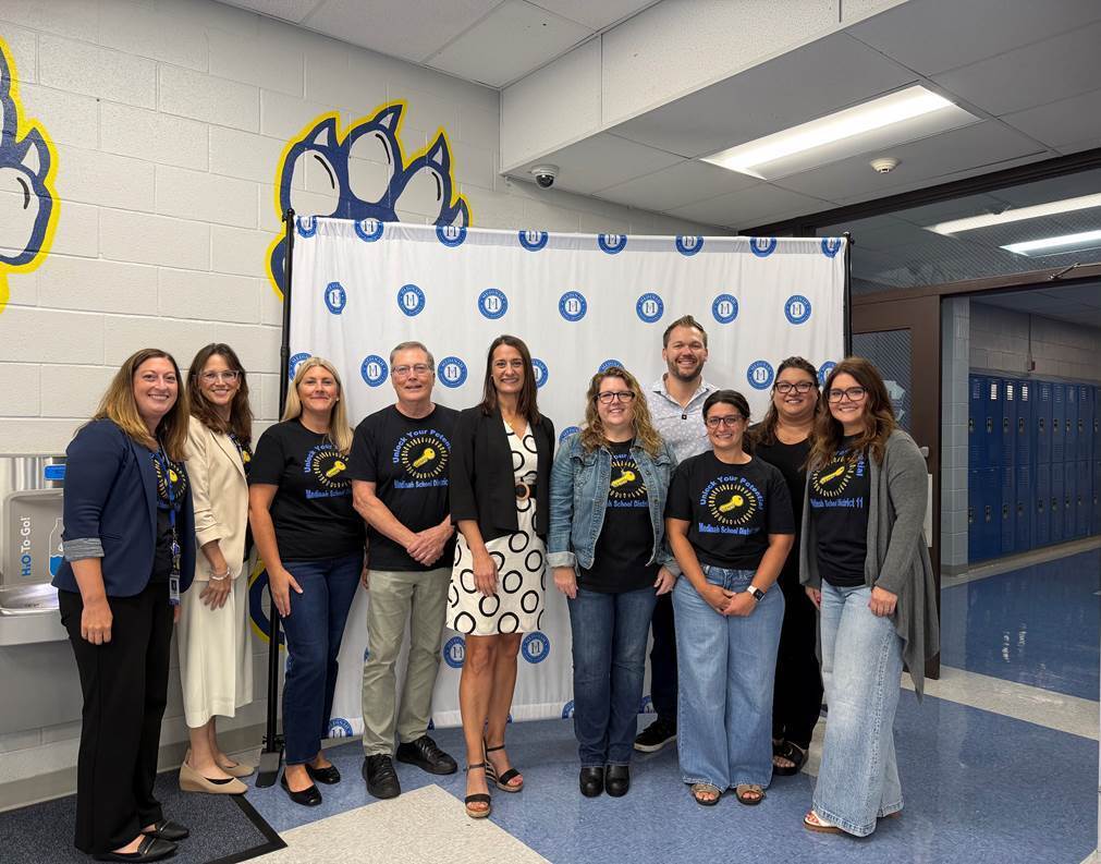 Eleven people pose for a photo in a school hallway with a colorful backdrop and a sink visible.