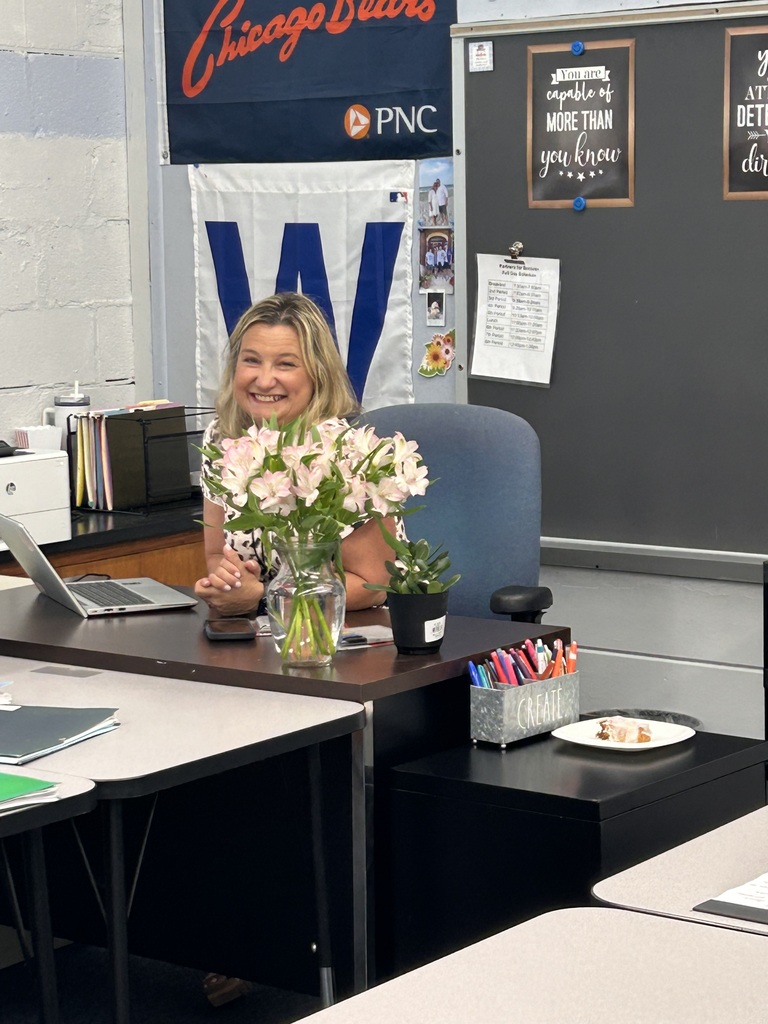A woman sits at a desk with a vase of flowers, a laptop, and other office items. Behind her, a whiteboard has text and pictures attached.