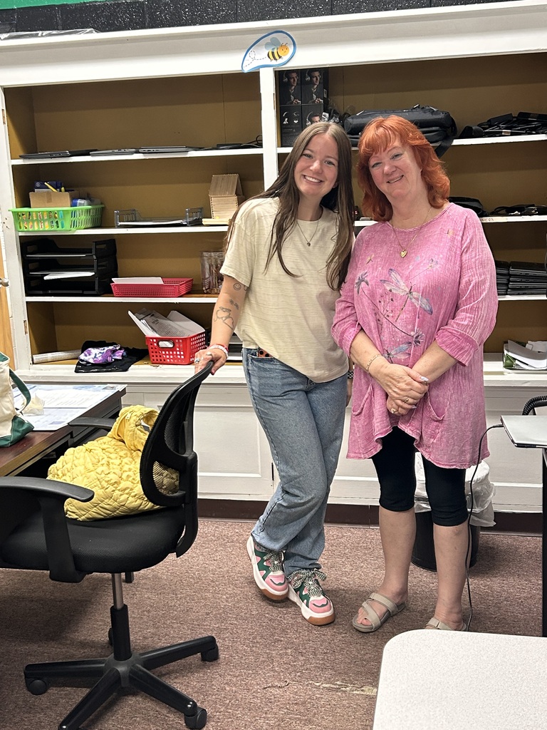 Two women in a room with shelves and a chair. One woman has a necklace, and the other has on sandals.