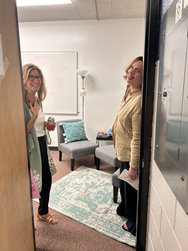 Two women, one with glasses, stand in an open doorway with a rug on the floor. Behind them, a chair, table, and lamp.